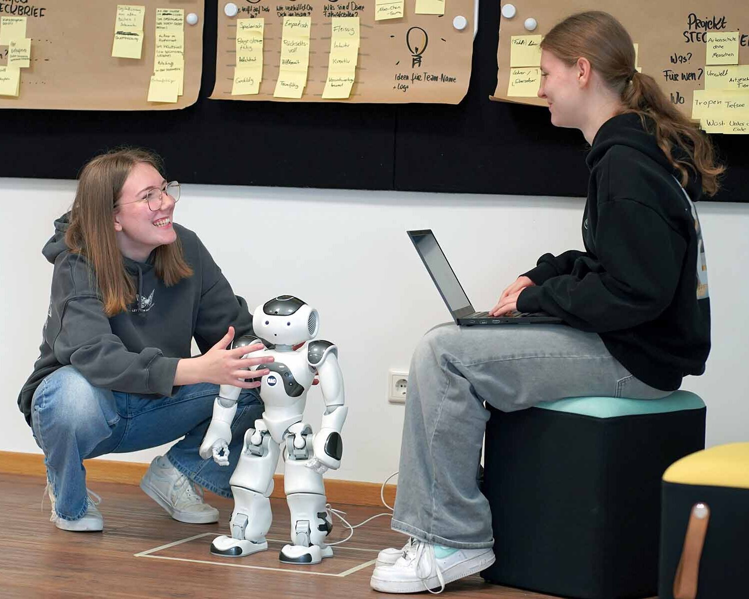 Ein Mädchen hockt neben Roboter, eines sitzt auf Hocker mit Laptop in der Hand