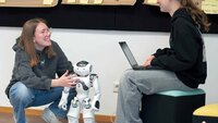 Ein Mädchen hockt neben Roboter, eines sitzt auf Hocker mit Laptop in der Hand