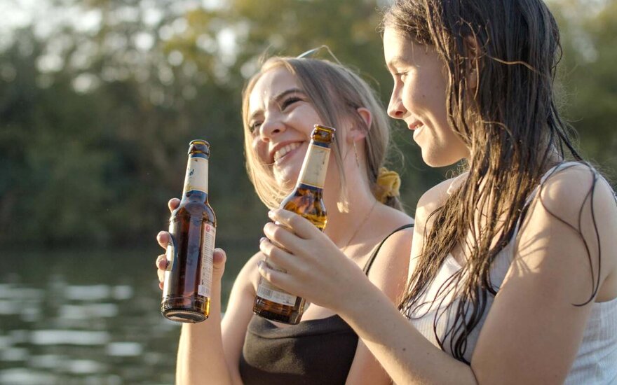 Zwei Studierende sitzen zusammen am Wasser, See, Fluss, bei Sonnenschein, lachen, trinken Bier, nasse Haare