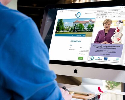 Mann in blauem Hemd sitzt vor einem Monitor, auf dem die Website des Information Management Instituts sowie das Cover der Studie mit Angela Merkel und einem Roboter zu sehen ist