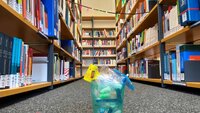 auf dem Boden vor mehreren Bücherregalen steht ein türkisfarbener Becher, der mit Ohrenstöpseln und einem Papierschirmchen gefüllt ist