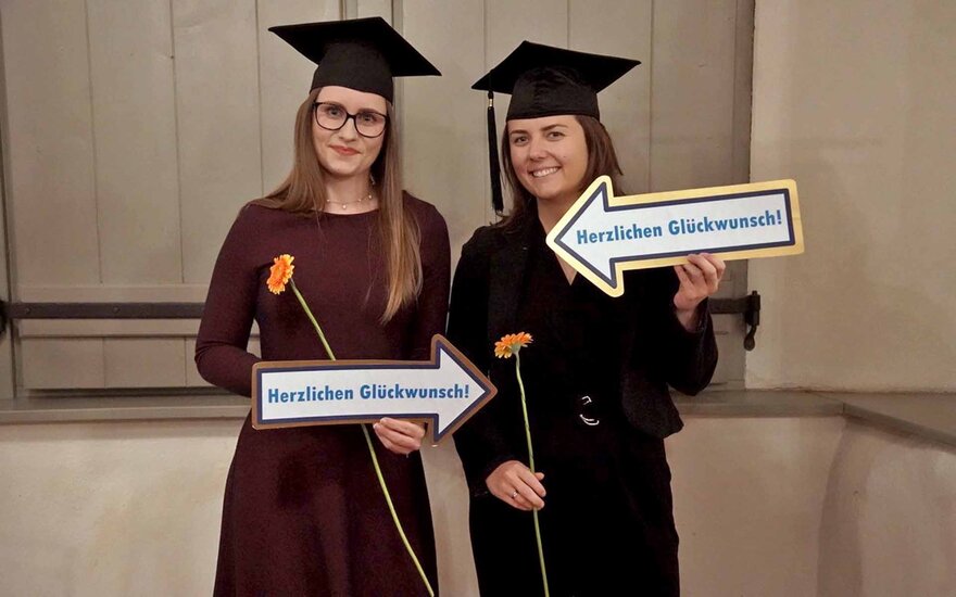 Zwei Studentinnen mit schwarzen Bachelorhüten, Rosen und Glückwunschschildern