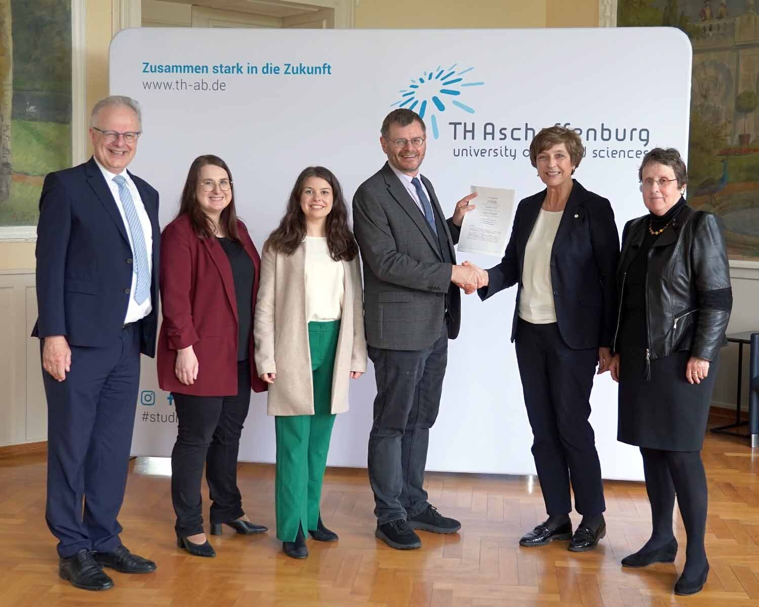 Gruppenbild mit zwei Männern und vier Frauen vor der TH-Logo-Wand im Hocksaal, Schulleiter und Präsidentin geben sich die Hand und halten den Vertrag