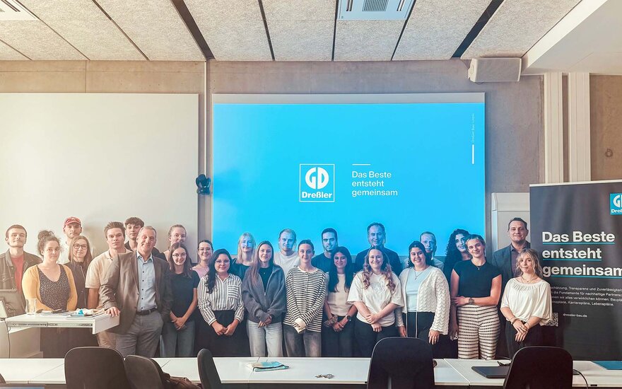 Gruppe von etwa 20 Personen steht in einem Seminarraum vor einer großen Projektionsfläche mit blauem Hintergrund und der Aufschrift „Das Beste entsteht gemeinsam“ sowie einem Firmenlogo von „Dressler Bau“. 