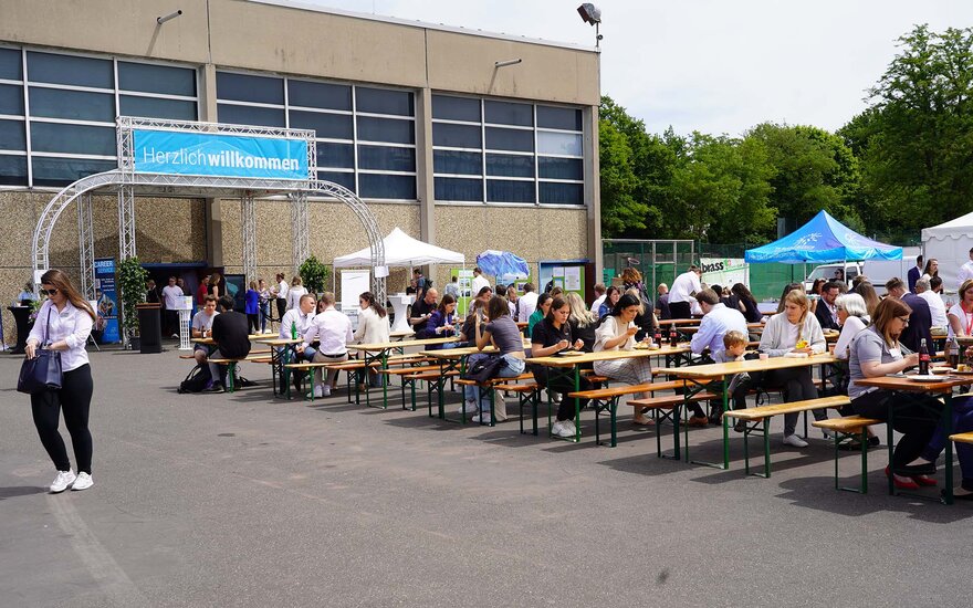 Zahlreiche Personen sitzen an Tischen im Freien, essen und sprechen miteinander. Im Hintergrund ist ein Gebäude zu sehen mit einem Banner, auf dem Herzlich willkommen steht