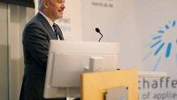 Professor Hofmann steht hinter einem Monitor am Rednerpult und spricht zum Publikum. Im Hintergrund ist eine Wand mit dem Logo der TH Aschaffenburg zu sehen.