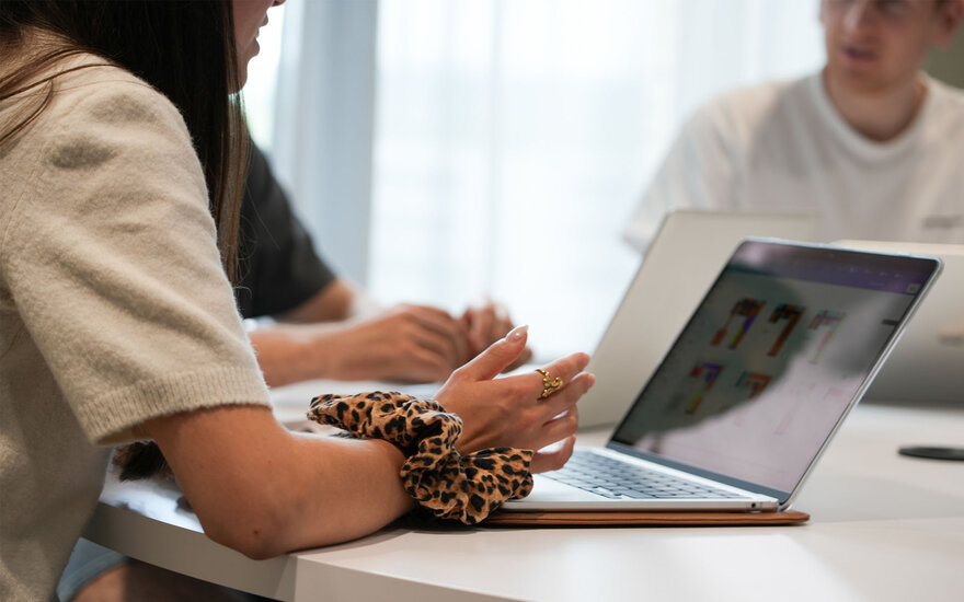 Studentin sitzt an einem Tisch und arbeitet an einem Laptop, auf dessen Bildschirm grafische Darstellungen zu sehen sind; im Hintergrund befinden sich weitere Personen mit Laptop.