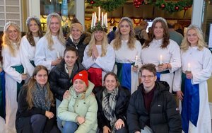 Fünf Studierende hocken mit Blick zur Kamera vor sieben jungen Frauen mit langen weißen Gewändern und silbernen Glitzerhaarbändern. Eine von ihnen trägt auf dem Kopf einen Kranz mit brennenden Kerzen.