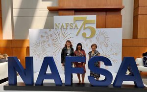 Drei Professorinnen stehen nebeneinander hinter dem NAFSA-Logo in blauen großen Buchstaben vor einer weißen Wand mit einem goldenen NAFSA-Logo und einer goldenen 75
