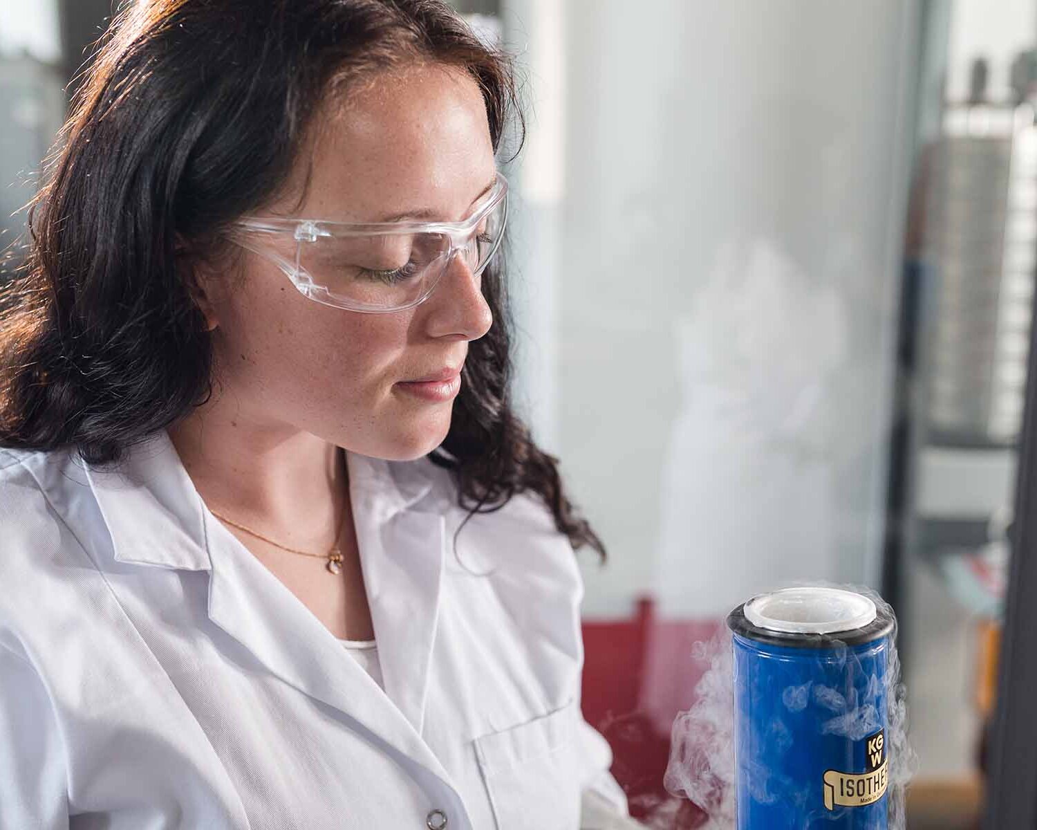 Studentin mit Schutzbrille und weißem Kittel hält ein Gefäß mit Stickstoff in der Hand
