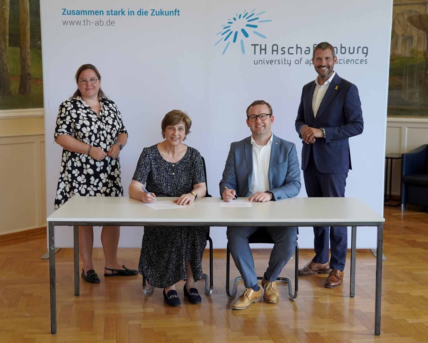 Präsidentin und BRK-Geschäftsführer sitzend bei der Vertragsunterzeichnung vor weißer Wand mit TH-Logo, Dekanin links und Landrat stehend rechts daneben