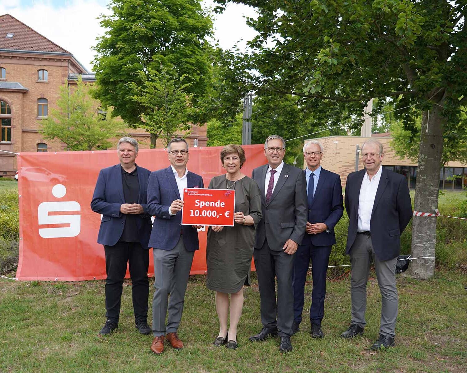 Präsidentin und Sparkassenvorstand mit rotem Schild mit Spendensumme in der Hand sowie Oberbürgermeister und drei Professoren in dunklen Anzügen stehen nebeneinander auf Campuswiese vor rotem Sparkassenbanner mit Backsteingebäude und Bäumen im Hintergrund