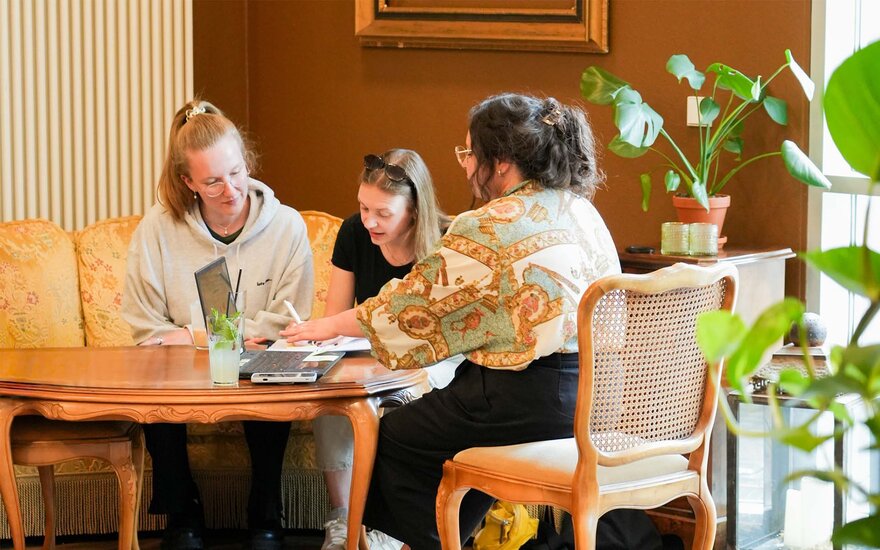 Drei Studentinnen sitzen gemeinsam an einem Holztisch im Campus-Café und arbeiten an einem Laptop. 