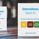 Zwei Tischaufsteller von Unternehmen an einem Messestand kommunizieren die Fachbereiche, in denen sie Studierende suchen.