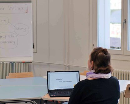 Eine Frau sitzt vor einem aufgeklappten Laptop mit Blick auf ein Flipchart