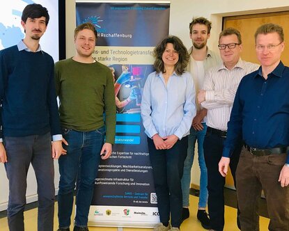 Die beiden Mitglieder des Clusters Sensorik stehen links neben den vier Vertreterinnen und Vertretern der TH Aschaffenburg vor einem Roll-up zum Wissens- und Technologietransfer