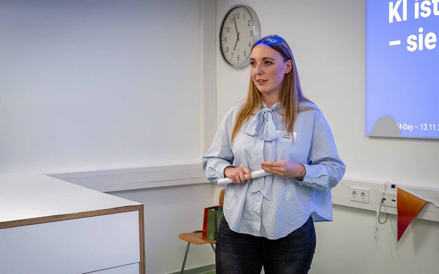 Frau mit langen blonden Haaren steht im Hörsaal am Rednerpult