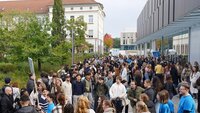 Große Menge an Studierenden zwischen einem modernen grauen Gebäude rechts und einem älteren weißen Gebäude mit rotem Dach und grünen Bäumen davor