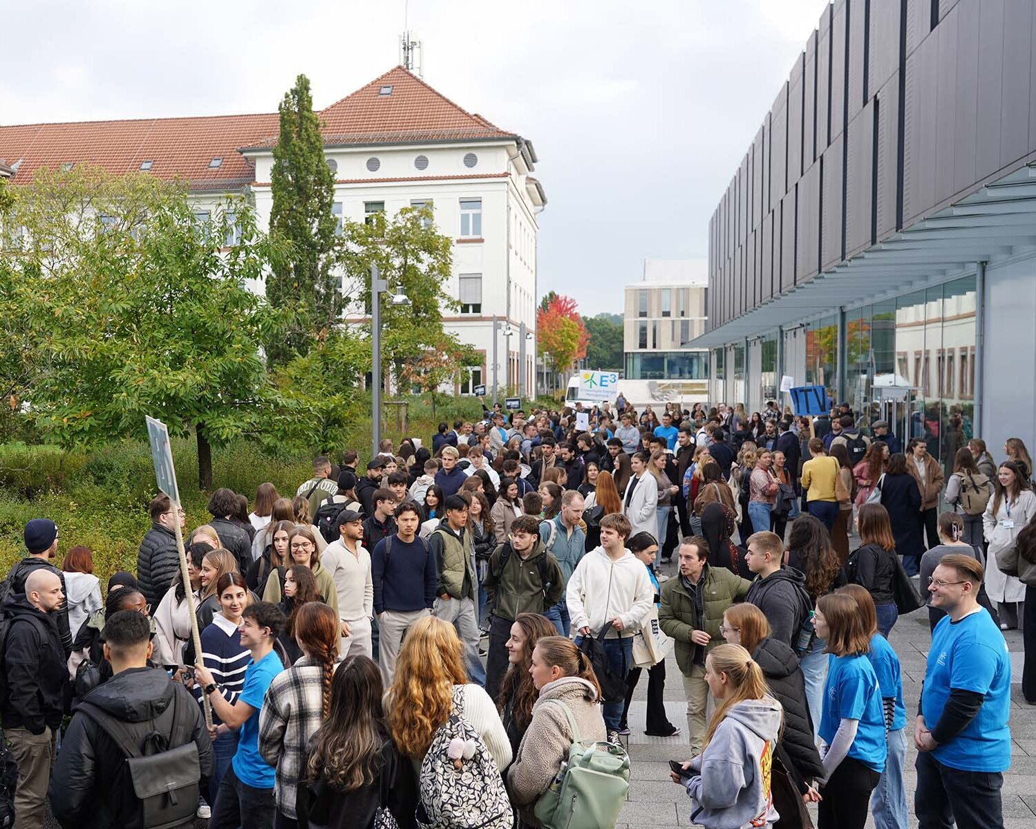 Große Menge an Studierenden zwischen einem modernen grauen Gebäude rechts und einem älteren weißen Gebäude mit rotem Dach und grünen Bäumen davor