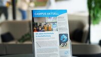 Hochschulmagazin im Prospektständer auf schwarzem Tisch im Foyer von Gebäude 48 mit beigem Sofa, grüner Pflanze und an der Scheibe vorbeilaufenden Personen im Hintergrund