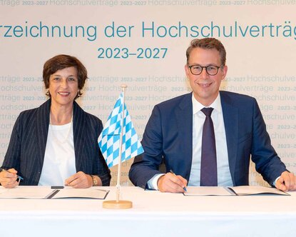 Die Hochschulpräsidentin sitzt links neben dem Wissenschaftsminister an einem Tisch und beide halten Stifte in der Hand, haben den Vertrag vor sich liegen und blicken in die Kamera