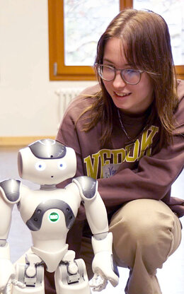 Mädchen schaut sich in der Hocke sitzend einen etwa 40 cm großen,  weißen Roboter an. Am anderen Ende des Raumes steht ein weiterer Roboter in der Unschärfe an einer Heizung.