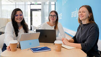 Drei Studierende sitzen an einem kleinen Tisch in einem modernen Aufenthaltsraum und arbeiten gemeinsam an Laptops.