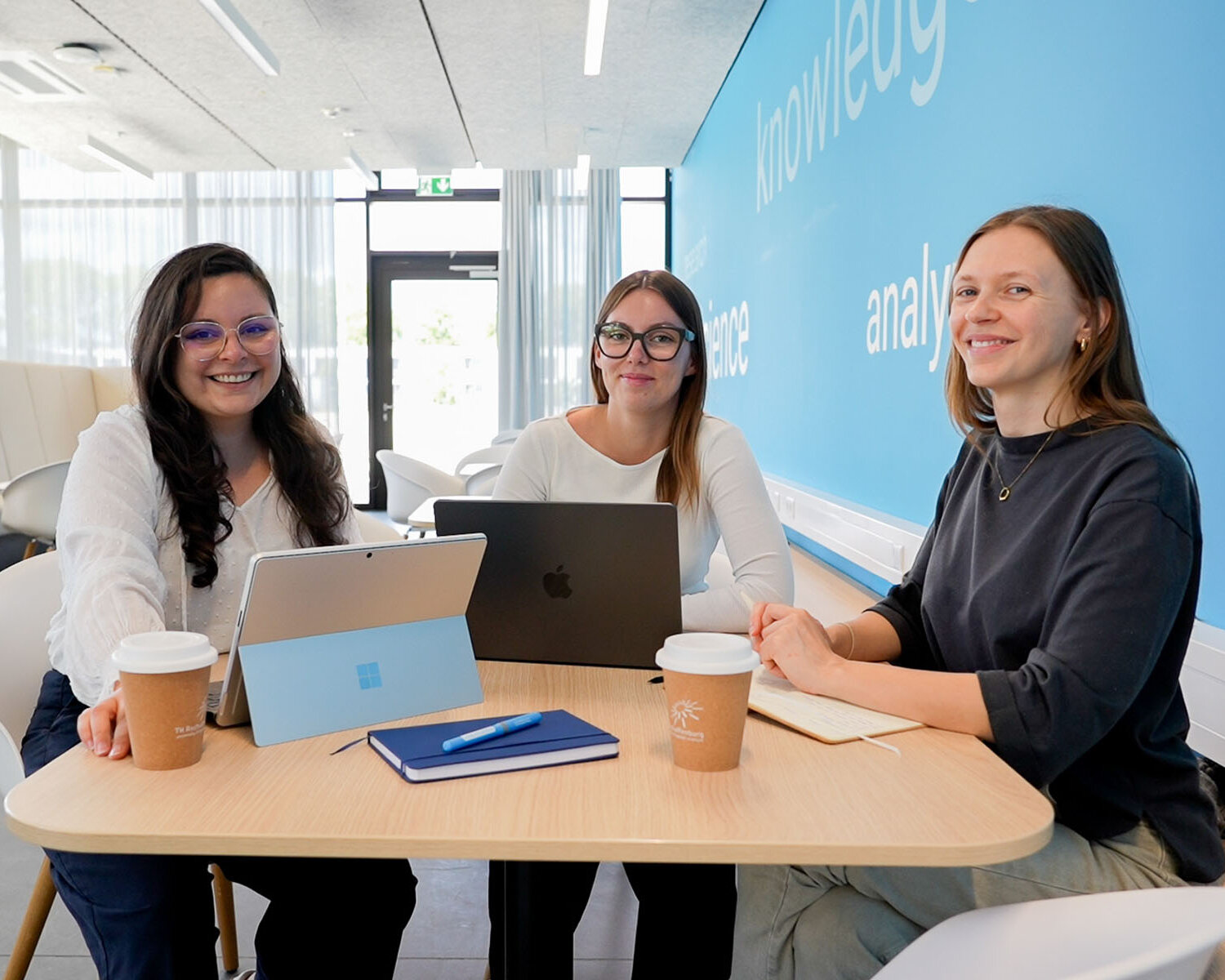 Drei Studierende sitzen an einem kleinen Tisch in einem modernen Aufenthaltsraum und arbeiten gemeinsam an Laptops.