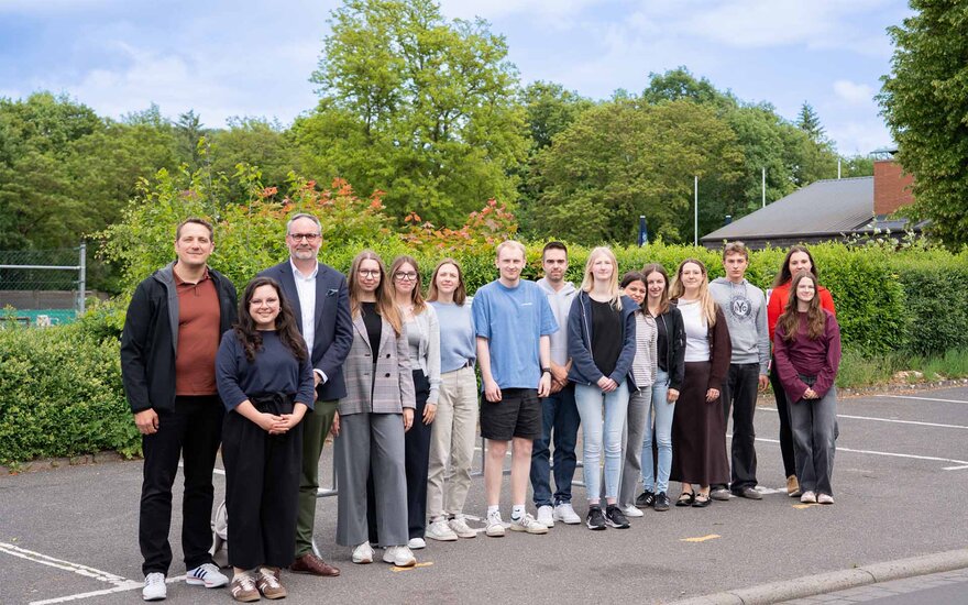 Gruppe von 15 Personen steht nebeneinander auf einem asphaltierten Platz im Außenbereich; im Hintergrund befinden sich grüne Bäume und Sträucher.