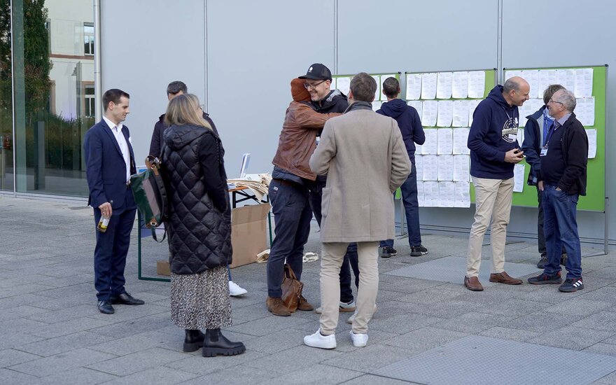 Mehrere Personen, die sich begrüßen und miteinander sprechen am Infostand vor der grauen Fassade des modernen Hochschulgebäudes Nr. 40