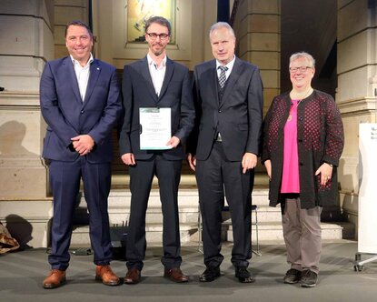 Der Preisträger hält lächelnd seine Urkunde in den Händen. Neben im in einer Reihe stehen Professor Mann, der IfKOM-Bundesvorsitzende Heinz Leymann und Cornelia Böhmer