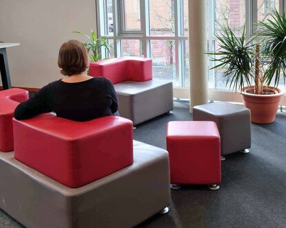 Ein rot-braunes Sofa, auf dem eine Frau in schwarzem Pullover sitzt und zum Fenster rausschaut, vor dem eine Topfpflanze steht