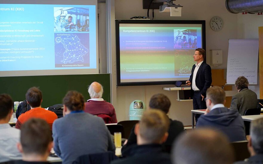 Ein Mann steht vor einer Leinwand und spricht zum sitzenden Publikum