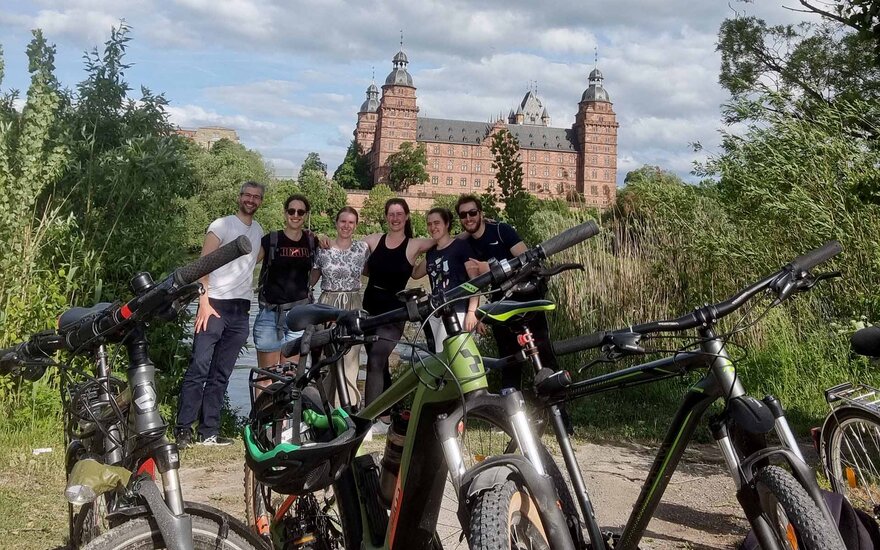 Eine Gruppe von Personen mit Fahrrädern im Vordergrund und dem Aschaffenburger Schloss im Hintergrund vor blauem Himmel