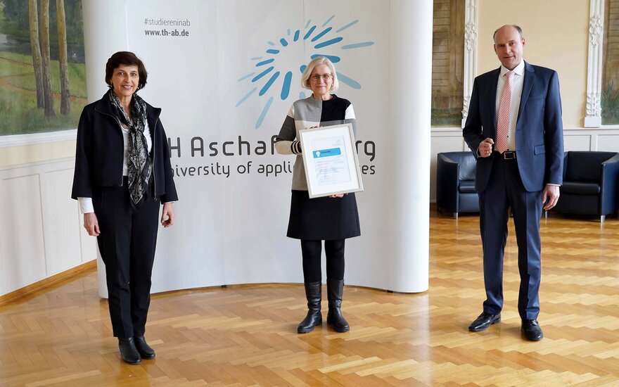 TH-Präsidentin, Schulleiterin mit der Urkunde in der Hand, und Professor stehen vor der Wand mit dem Logo der TH Aschaffenburg im Hock-Saal