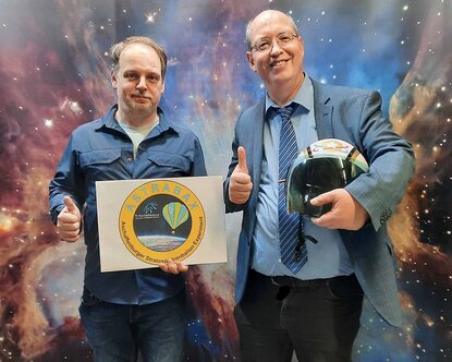 Professor Hildenbrand hält das Logo des Projekts in der Hand, Professor Döhring einen Astronautenhelm. Beide heben den Daumen der rechten Hand in die Höhe und stehen vor einer Wand, die einen Sternenhimmel zeigt