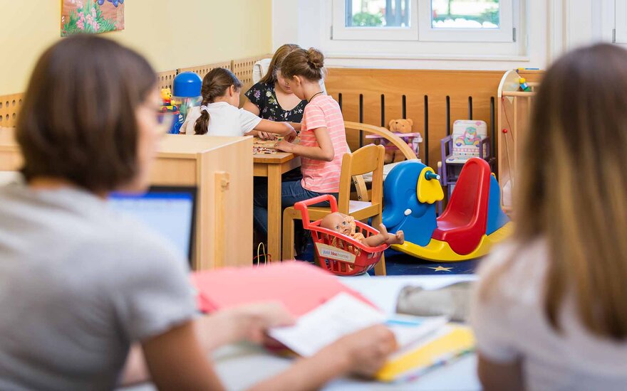 Zwei junge Frauen sitzen am Tisch vor spielenden Kindern im Hintergrund