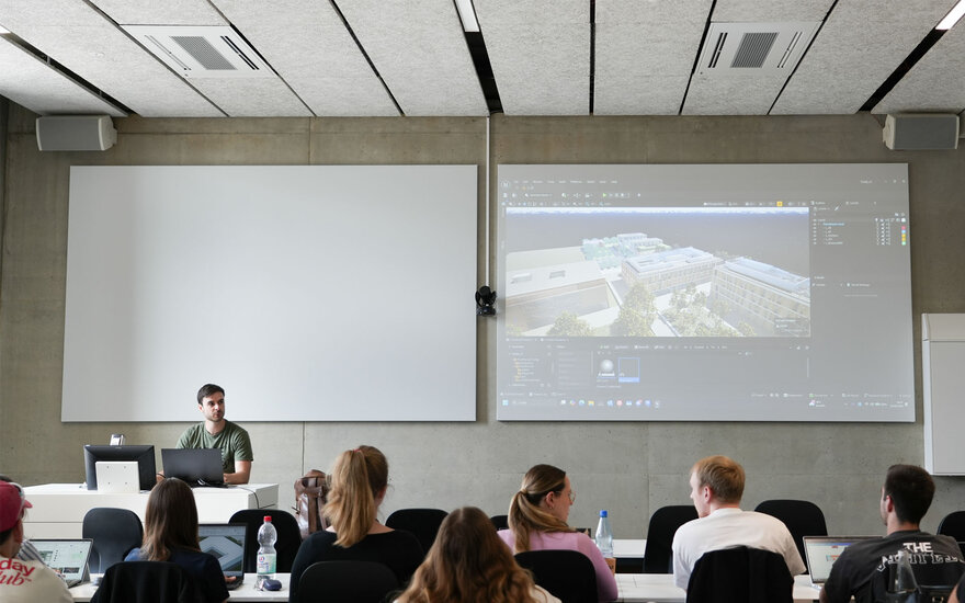 Vorlesungsraum mit mehreren Studierenden, die an Tischen sitzen. Auf einer großen Leinwand ist eine 3D-Visualisierung eines Gebäudekomplexes zu sehen.