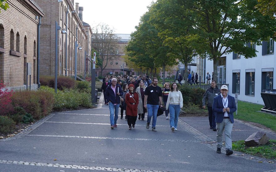 Gruppe von rund 50 Personen, die von einem Mann mit Kappe, Brille und blauem Jacket über den asphaltierten Campusweg zwischen einem älteren und einen modernen Gebäude geführt werden