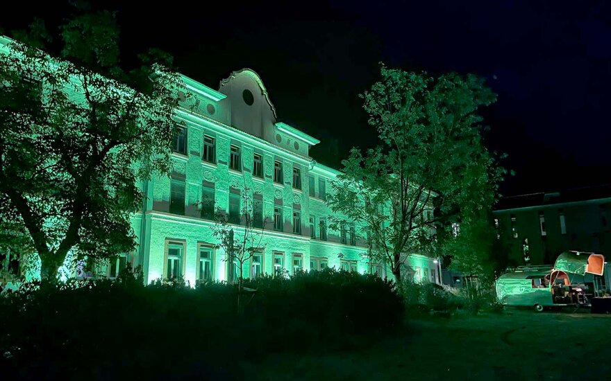 Grün beleuchtete Fassade von weißen Hochschulgebäude mit zwei Bäumen im Vordergrund