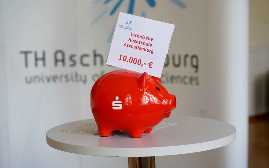 rotes Sparschwein mit Zettel der Spendensumme von 10.000 Euro steht auf einem runden Tisch vor dem Logo der TH Aschaffenburg