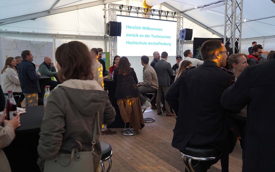 Alumni stehen und sitzen im Zelt und unterhalten sich, im Hintergrund weiße Leinwand mit der blauen Aufschrift "Herzlich willkommen zurück an der Technischen Hochschule Aschaffenburg"