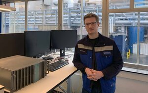 Ein junger Mann mit Brille im blauen Arbeitsanzug lehnt an einem Tisch mit Monitoren und technischen Geräten im Hintergrund