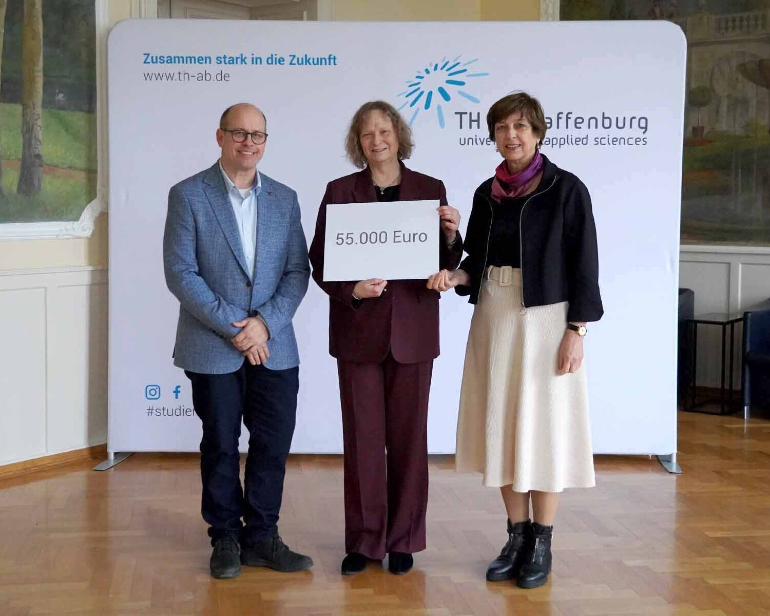 Frau in der Mitte hält Schild mit Spendensumme, Mann und Frau stehen rechts und links neben ihr vor TH-Logowand