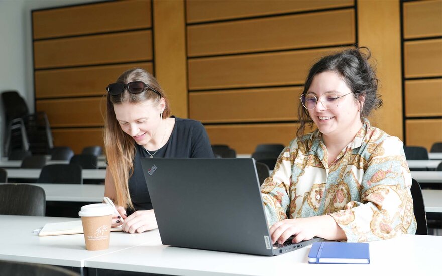 Zwei Studierende sitzen in einem Seminar an einem Tisch und arbeiten an einem Laptop und handschriftlichen Notizen; auf dem Tisch stehen ein Kaffeebecher und ein Notizbuch. 
