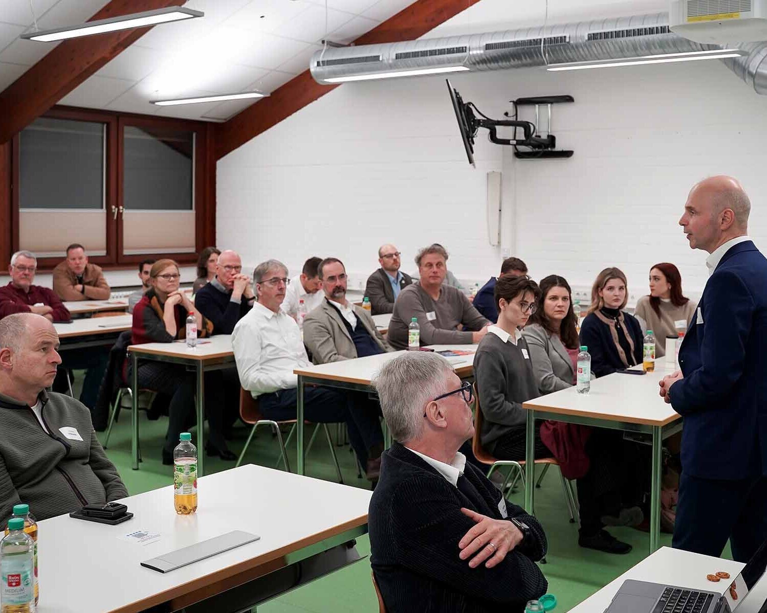 Mann im schwarzen Anzug steht im Hörsaal vor Teilnehmenden an Tischen und hält einen Vortrag