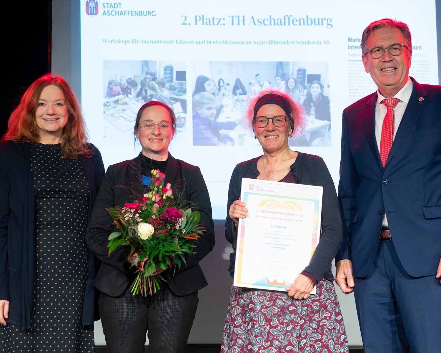 Gruppenfoto von der Urkundenverleihung mit drei Frauen, eine davon mit Urkunde, eine mit Blumenstrauß und einem Mann