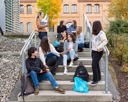 Ein Gruppe von internationalen Studierenden sitzt auf einer Treppe an der Hochschule vor einem gelbbraunen Backsteingebäude und ist im Austausch miteinander