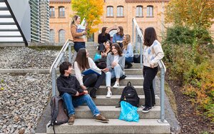 Ein Gruppe von internationalen Studierenden sitzt auf einer Treppe an der Hochschule vor einem gelbbraunen Backsteingebäude und ist im Austausch miteinander