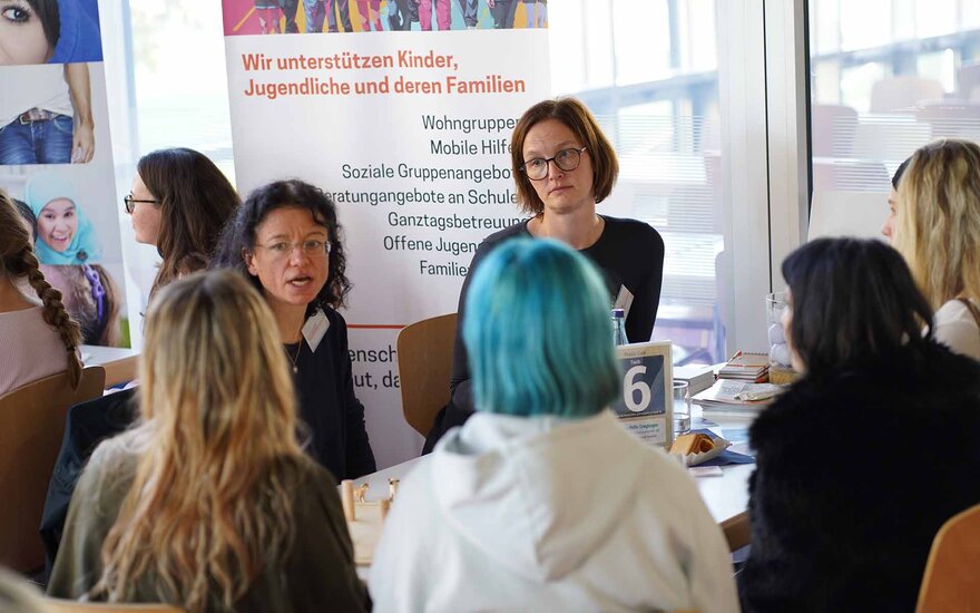 Zwei Sozialarbeiterinnen unterhalten sich am Tisch sitzend mit vier Studentinnen